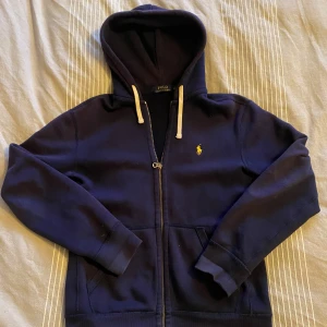 Polo Ralph Lauren zip up - Säljer en snygg mörkblå hoodie från Polo Ralph Lauren med dragkedja och det klassiska broderade logotypen i gult på bröstet. Tröjan har långa ärmar och en bekväm huva med snörning. Perfekt för en avslappnad stil.