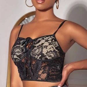 Svart spetskorsett med knytning - Trendig svart spetskorsett med justerbara axelband och knytning framtill. Den har ett vackert blommönster och är perfekt för en stilfull look. Passar bra till både jeans och kjol.