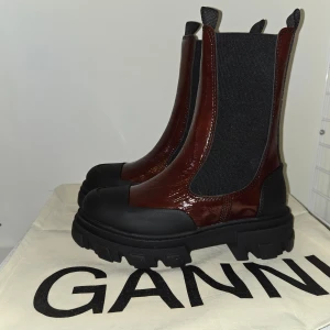 Burgundy boots från Ganni - Snygga Burgundy Chelsea boots från Ganni med hög sula och elastiska paneler för enkel påtagning. Skorna har en robust design med svarta detaljer och är perfekta för en trendig look. Är små I storlek, är storlek 38 men passar nog bättre en 37. Aldrig använda, fått i julklapp men för små tyvärr.