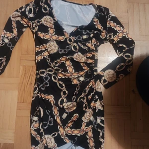 Svart klänning med kedjemönster - Snygg svart klänning med ett iögonfallande kedjemönster i guld och beige. Klänningen har en v-ringad hals och långa ärmar, vilket ger en elegant look. Perfekt för att sticka ut på festen!