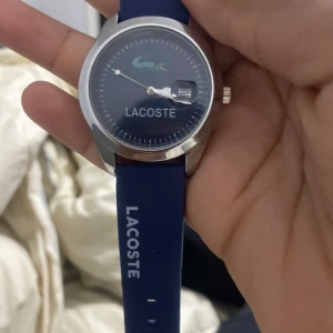 Blå klocka från Lacoste - Snygg blå klocka från Lacoste med en stilren urtavla och datumvisning. Klockan har ett silverfärgat hölje och ett matchande blått armband med Lacoste-loggan. Perfekt för en sportig och modern look.