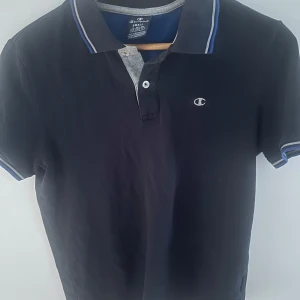 Mörkblå pikétröja från Champion - Snygg mörkblå pikétröja från Champion med blå och grå detaljer på kragen. Tröjan har korta ärmar och en liten logga på bröstet. Perfekt för en avslappnad stil.