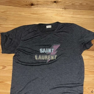 Grå t-shirt från Saint Laurent - Snygg grå t-shirt från Saint Laurent med ett coolt blixtmotiv och texten 'SAINT LAURENT' i pastellfärger. Perfekt för en avslappnad stil med en touch av lyx. Storlek m men passar även S, skriv privat för frågor 