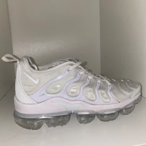 Vita Nike Air Vapormax Plus - Snygga vita Nike Air Vapormax Plus med en futuristisk design och transparent sula. Skorna har snörning och en bekväm passform, perfekt för en modern look.