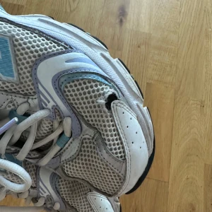 Vita sneakers från New Balance - Säljer ett par vita sneakers från New Balance med ljusblå och lila detaljer. Skorna har en luftig mesh-design och klassisk snörning. Perfekta för en sportig look. Tyvärr är de trasiga i häl och på sidorna. Har satt hälskydd i skorna men den händiga kan säkert laga skadorna på sidan🩵
