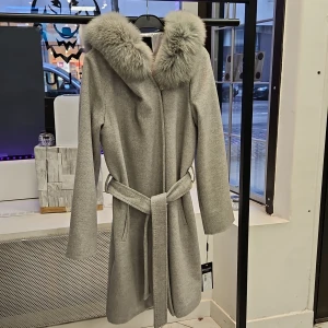 Grå ull kappa Lucinda från Hollies - Elegant grå ull kappa från Hollies med en lyxig pälskrage och knytbälte i midjan. Perfekt för kyliga dagar med sin stilrena design och långa ärmar. Kappan har en klassisk look som passar till många tillfällen. Kappan är mindre I storleken, motsvarar en 38 storlek. Material: Ull och pälskrage är äkta. Ny med etikett.