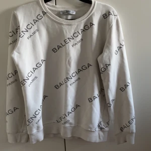 Vit sweatshirt från Balenciaga - Säljer en vit sweatshirt från Balenciaga med svart logotyptryck över hela tröjan. Den har långa ärmar och en rund halsringning. Perfekt för en stilren och modern look. ((( som ni ser på 4de bilden är där en jätte jätte lite fläck med den syns knappt)))
