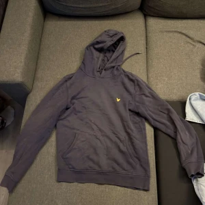Mörkblå hoodie från Lyle & Scott - Säljer en mörkblå hoodie från Lyle & Scott med en gul logotyp på bröstet. Tröjan har en klassisk känguruficka och en bekväm huva med dragsko. Perfekt för en avslappnad stil.  Har fått blekning på sig vilket gör att den missfärgats, se bild.