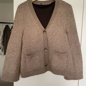 Beige/brun stickad kofta med knappar - Säljer en mysig stickad kofta med stora knappar framtill. Koftan har två praktiska fickor och ribbade muddar vid ärmslut och nederkant. Perfekt för kyliga dagar!
