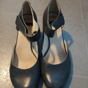 Svarta klackskor från Vagabond - Snygga svarta klackskor från Vagabond i skinn med en bekväm rem över foten. Skorna har en stabil klack och rund tå, vilket gör dem både stiliga och bekväma.