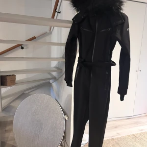 Svart overall med pälsdetaljer från ASOS 4505 - Snygg svart overall från ASOS 4505 i storlek 34 med pälsdetaljer på huvan. Den har dragkedja framtill och justerbart bälte i midjan för en perfekt passform. Perfekt för vinterns äventyr i snön! ❄️köpte den nyss men storleken  passade inte så säljer den vidare. 🎿