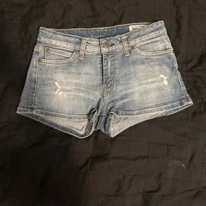 Blå jeansshorts - Snygga blå jeansshorts med slitna detaljer och klassisk femficksdesign. Perfekta för en avslappnad stil. De har en knapp och dragkedja framtill samt bälteshällor.