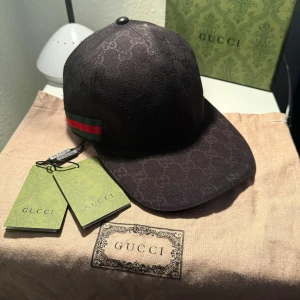 Gucci Canvas Keps - Snygg svart keps från Gucci med det ikoniska GG-mönstret. Kepsen har en klassisk design med en röd och grön randdetalj på sidan. Perfekt för att ge en lyxig touch till din outfit.