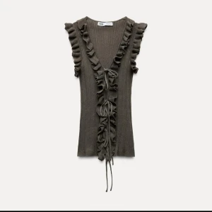 Populära volangtopp från Zara  - Snygg khakigrön ribbad topp från Zara med volangdetaljer längs den v-ringade halsen. Toppen är ärmlös och har knytband framtill. Perfekt för en stilren look. Oanvänd med prislapp 