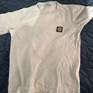 Vit t-shirt från Stone Island - Säljer en stilren vit t-shirt från Stone Island med korta ärmar. Den har en broderad logotyp på bröstet. Perfekt för en avslappnad look. Nypris ligger på 1000kr ish. Köpt på Nk i Stockholm 
