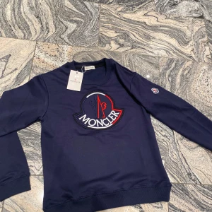 Mörkblå tröja från Moncler - Säljer en snygg mörkblå tröja från Moncler med ett stort broderat logotypmärke på bröstet och ett mindre på ärmen. Tröjan har långa ärmar och ribbade muddar vid ärmslut och nederkant. Perfekt för en stilren look!