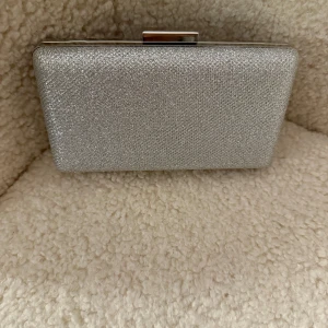 Glittrig silver kuvertväska - Snygg och glittrig kuvertväska i silver med en elegant kedja för axelrem. Perfekt för att ge en touch av glamour till din outfit. Väskan har en stilren design med en praktisk stängning upptill.