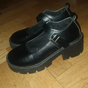Svarta chunky Mary Jane skor - Säljer ett par svarta Mary Jane skor med chunky sula och justerbart spänne. Skorna har en rund tå och är tillverkade i syntetmaterial. Perfekta för en edgy look.
