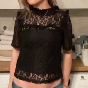 Svart spetsblus - Säljer en elegant svart spetsblus med korta ärmar och hög hals. Blusen har ett genombrutet mönster och är perfekt för en stilfull look. Passar bra till både jeans och kjol. Strl S/32 men passar mig som vanligtvis bär M