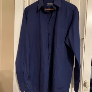 Blå skjorta från Eterna - Snygg blå skjorta från Eterna med klassisk krage och knappar framtill. Skjortan har en diskret broderad detalj på bröstfickan. Perfekt för en stilren look.