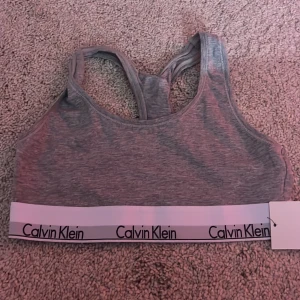 Grå bralette från Calvin Klein - Säljer en snygg grå bralette från Calvin Klein med elastiskt band och logga. Den är tillverkad i mjukt material och har en sportig design. Aldrig använd. Väldigt mjukt material. Storlek S/M. Pris kan diskuteras. 