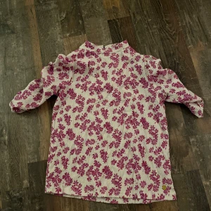 Blommig lila topp med puffärmar - Lila/rosa topp med blommönster med en ljuslila bakgrund. Den har puffiga trekvartsärmar och en hög krage. Köpt på gekås, Pernilla Wahlgrens märke. Använt några gånger men bra skick, klippt bort lappen men storlek S/M typ. Om du har frågor eller vill ha mer bilder kontakta mig🩷