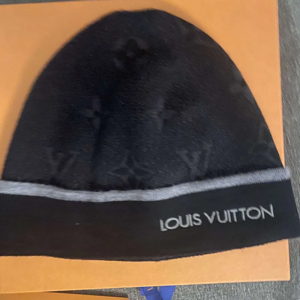 Elegant svart mössa från Louis Vuitton med subtilt mönster och grå randdetalj. Märkesnamnet är broderat i vitt på kanten, vilket ger en lyxig touch. Perfekt för att hålla stilen under kyliga dagar.. Asusteet.