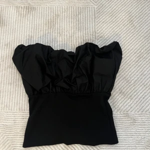 Svart axelbandslös topp - Snygg svart axelbandlös topp med voluminös design upptill. Perfekt för en stilren look, tillverkad i ett mjukt material som ger en bekväm passform. Passar utmärkt till både jeans och kjol. Endast använt en gång.