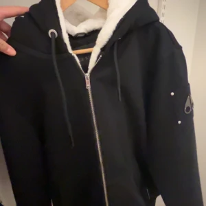 Svart jacka med dragkedja från Moose Knuckles - Säljer en svart jacka från Moose Knuckles med dragkedja och huva. Jackan har en snygg detalj på ärmen och är fodrad med mjukt material för extra värme. Perfekt för kyligare dagar!