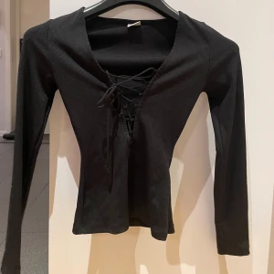 Svart ribbad topp från Gina Tricot - Säljer en svart ribbad topp från Gina Tricot i storlek XS. Toppen har långa ärmar och en snygg snörning framtill som ger en v-ringad look. Perfekt för en stilren och trendig outfit.