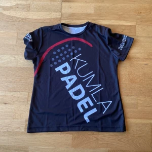 Svart padel tränings tröja med tryck  - Snygg svart t-shirt från Kumla Padel med stort tryck på framsidan och flera logotyper på baksidan. Den har korta ärmar och en rund halsringning. Perfekt för padelentusiaster!