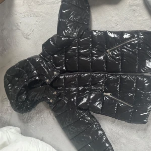 Svart glansig jacka  från Nelly - Snygg svart pufferjacka från Nelly med glansig finish och dragkedja framtill. Jackan har en huva och är perfekt för kyligare dagar. Den quiltade designen ger en modern look.