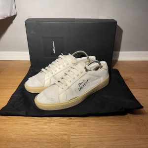 Vita sneakers från Saint Laurent - Snygga vita sneakers från Saint Laurent med klassisk design och märkets logga på sidan. Skorna har en stilren men har tyvärr lossnat lite kring sulan. Har försökt limma men gav ingen jätte kärlek till de. Enkelt att fixa själv! Annars inga konstigheter och hör av dig om du har frågor eller om du är intresserad av ett byte. Tveka inte om du har frågor eller vill se fler bilder!