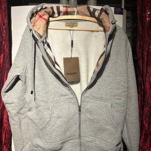 Grå hoodie från Burberry - Säljer en stilren grå hoodie från Burberry med klassiskt rutigt mönster i huvan. Tröjan har dragkedja framtill och en broderad logga på bröstet. Perfekt för en avslappnad och trendig look. OBS! Priset är inte hugget i sten. Hör av er vid minsta fundering 🍾