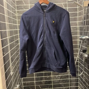Mörkblå hoodie från Lyle & Scott - Säljer en snygg mörkblå hoodie från Lyle & Scott med dragkedja och huva. Den har ett broderat gult emblem på bröstet och är perfekt för en avslappnad stil. Passar ungdomar i åldern 14-15 år.