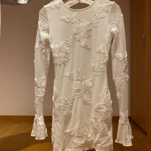 Blommig student klänning med öppen rygg - Säljer en elegant vit klänning med blommigt mönster och öppen rygg. Klänningen har långa ärmar med volangdetaljer vid ärmsluten. Perfekt som student klänning! Klänningen är endast använd 1 gång. 