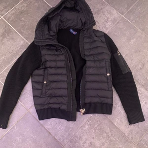 Svart jacka från Moncler - Snygg svart jacka från Moncler med huva och dragkedja. Jackan har en quiltad design med ribbade ärmar och muddar. Perfekt för kyligare dagar. Tyvärr är den sönder lite på ena mudden, men inget som syns. 