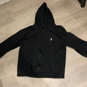 Svart huvtröja från Ralph Lauren - Säljer en stilren svart huvtröja från Ralph Lauren med dragkedja och broderad logga på bröstet. Tröjan har långa ärmar och en praktisk huva. Perfekt för en avslappnad look.