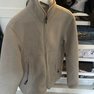 Beige fleecejacka med dragkedja - Säljer en mysig beige fleecejacka med dragkedja. Jackan har en hög krage och två praktiska fickor med dragkedjor. Perfekt för kyliga dagar när du vill hålla dig varm och bekväm. Samt att denna är knappt använd och att skicket är 10/10 , märke Arket 