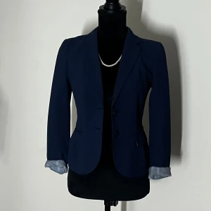 Mörkblå kavaj - Elegant mörkblå kavaj med två knappar framtill och klassisk krage. Kavajen har en figursydd passform och långa ärmar med ljusare foder som kan vikas upp för en stilfull detalj. Perfekt för en sofistikerad look.