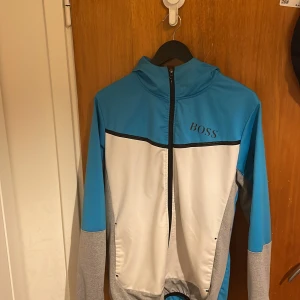 Blå och vit jacka från Boss - Säljer en snygg blå och vit jacka från Boss med dragkedja framtill. Jackan har en sportig look med gråa ärmar och en hög krage. Perfekt för en casual stil.