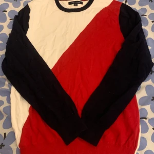 Tommy Hilfiger  - Säljer en snygg flerfärgad tröja från Tommy Hilfiger i storlek M. Tröjan har ett diagonalt mönster i vitt, rött och blått med långa ärmar. Perfekt för en stilren look.