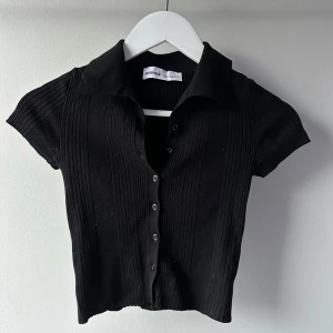 Svart ribbad kortärmad topp från Bershka - Snygg svart ribbad topp från Bershka med korta ärmar och knappar framtill. Perfekt för en stilren look. Toppen har en klassisk krage och är i en tight passform.
