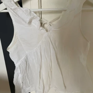 Vit ärmlös topp från Gina Tricot - Säljer en söt vit ärmlös topp från Gina Tricot. Toppen har volangdetaljer vid axlarna och en knytning i ryggen. Perfekt för varma sommardagar. Passar både till jeans och kjol!
