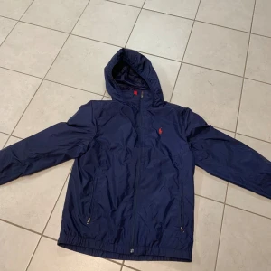 Mörkblå vindjacka från Polo Ralph Lauren - Snygg mörkblå vindjacka från Polo Ralph Lauren med huva och dragkedja. Jackan har långa ärmar och två fickor med dragkedja framtill. Perfekt för blåsiga dagar.