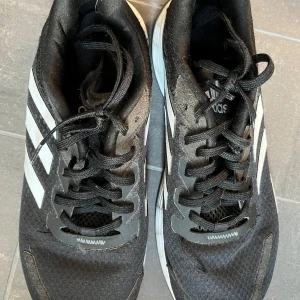 Svarta Adidas sneakers med vita detaljer - Snygga svarta Adidas sneakers med klassiska vita ränder och snörning. Skorna har en robust sula med bra grepp och Adidas-logga på undersidan. Perfekta för en sportig look.