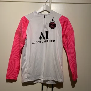 Vit och rosa fotbollströja från Jordan x PSG - Snygg vit fotbollströja med rosa mönstrade ärmar från Jordan x PSG. Tröjan har en halv dragkedja framtill och svart text med 'Accor Live Limitless' på bröstet. PSG-loggan och Jordan-märket syns tydligt. Perfekt för fotbollsfans som vill sticka ut!