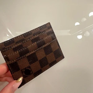 Brun och svart rutig korthållare  - Snygg och kompakt plånbok i brunt och svart med ett klassiskt rutigt mönster. Perfekt för att hålla dina kort organiserade. Den är stilren och lätt att bära med sig.