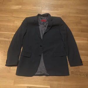 Grå kavaj från Hugo Boss - Snygg grå kavaj från Hugo Boss med klassisk design. Kavajen har två knappar framtill och ett elegant innerfoder. Perfekt för en stilren look. En lapp saknas på baksidan av ena ärmen därav det bra priset (se bild). Nypris på denna går på 4000 kr Bröstmått: 58 cm  Axlar 47 cm Ärmlängd 69 cm 