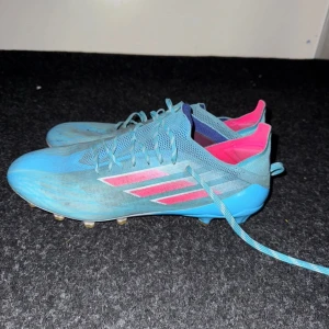 Adidas Speedflow  - Säljer ett par blå fotbollsskor från Adidas med rosa detaljer och snörning. Skorna har dobbar för bra grepp på planen och en modern design med meshmaterial för ventilation.